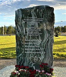 An upright stone slate with flowers placed at its base. On the slate there are two blocks of text, one in Norwegian, and the other in English. The English text reads as follows: "In memory of our irreplaceable youths, who lost their lives in the helicopter accident in the Skoddevarre mountain August 31, 2019". Below the English text, there is a list of names and ages. At the very bottom of the slate, there is an outline of a heart