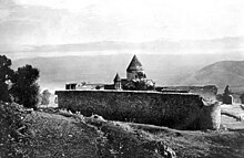 A photograph of the now-destroyed Arakelots Monastery showing a stone wall and the tops of several buildings behind it.