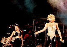 Madonna, dressed in a bone colored corset with black pants, stretches out her left arm. To her left, there is a brunette female wearing a black bra and pants. Behind the two females, several drums and smoke can be seen.