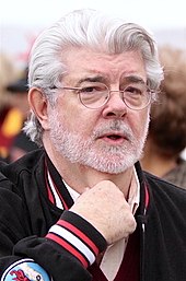 George Lucas sporting white hair and a short beard looking at the camera