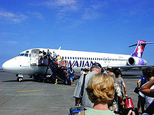 People line up to board a white twin-engine plane on a sunny day