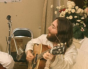 A man with long dark hair and a beard, wearing glasses and a white jacket, playing a guitar