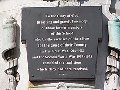 Affixed to the weathered, discoloured white stone war memorial is a black plaque with white text in Times New Roman font. The text, which is centred and has generous line spacing, reads: "To the Glory of God / In lasting and grateful memory / of those former members / of this School / who by the sacrifice of their lives / for the cause of their Country / in the Great War 1914 – 1918 / and the Second World War 1939 – 1945 / ennobled the traditions / which they had here received."