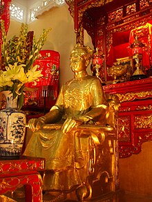 A golden-coloured statue of a man in a gown on a seat with a sword on his knees. In front there is a polished wooden table with goldleaf and a blue and white porcelain vase with yellow flowers. Behind him is a wooden altar with lights and incense holders. The altar has the same design as the table. The wall is cream-coloured.