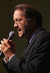 A middle-aged man wearing glasses and a dark suit, talks to a microphone.