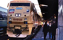 A diesel locomotive in tan paint. Lettering includes "Amtrak" and "Celebrate the Century Express". A United States Postal Service logo and a stamp logo reading "100 Celebrate the Century" are on the front.