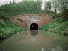 Red brick tunnel entrance to a tunnel through which light can just be seen at the far end. On either side are grassy banks down to the water.