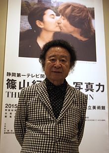 A Japanese man standing behind a poster-board, featuring a photograph of John Lennon and Yoko Ono kissing in New York.