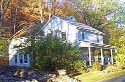 A white house with a perpendicular rear wing. There is a large green shrub in front, pavement in the lower right corner and trees, some of which have turned orange with fall, in the rear.