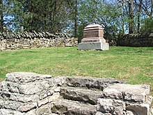 A single historical marker in a grassy area that sits in a flat raised area accessible by five roughly carved limestone stairs. The area is enclosed with low walls of stacked flat slabs of limestone. Pine and other trees surround the area outside the stone enclosure. The grave is marked with about four large, carved rectangular blocks of stone stacked with the largest gray stone at the bottom, and polished shiny pink stones on top. One stone has in raised carved letters "LEATHERLIPS", and above that is a stone with this carved inscription: "LEATHERLIPS, a chief of the Wyandot Tribe of Indians was executed on this spot June 1, 1810. Erected by the Wyandot Club of Columbus Ohio 1889."