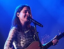 In blue lighting especially visible on her hair, a woman performs onstage with an acoustic guitar and a big smile.