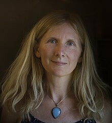 Portrait of Samantha Harvey, a middle-aged woman with blonde hair and light-coloured eyes, wearing a blue necklace and sundress.