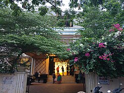 A building entrance, flanked by trees, revealing a lighted doorway above a short flight of stairs, with people standing in front..