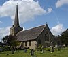 A long, low grey stone church with a tiled roof and a very tall spire at its far end.