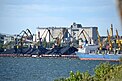 Am Pier liegt ein Frachtschiff mit geöffneten Luken. Das Bild zeigt sechs Hafenkräne, Eisenbahnwagen und Erzlager vor großen Silos im Hintergrund.