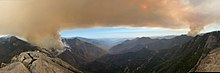 Two plumes of smoke rise on either side of a deep, forested canyon in a panoramic shot