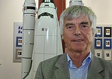 Ulf Merbold with gray hair in front of a model of the Space Shuttle