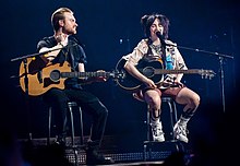 Billie Eilish and Finneas O'Connell seated at center stage, holding their acoustic guitars