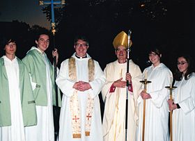 両脇の4名はアコライト（侍者）。中央左側の司祭はアルブと白のストールを着用し、中央右側の主教は白のチャジブルと金色のマイター（主教帽）を身に着け、牧杖（パストラル・スタッフ）を持っている。