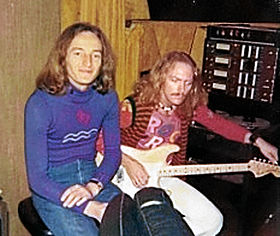 Two musicians sit side by side in a recording studio. Both men have long red hair. The man on the left wears a blue jumper and sits in a relaxed manner. The one on the right is tuning his guitar.