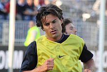 Portrait d'un joueur de football. Il porte au-dessus de son maillot noir une chasuble jaune avec le logo de l'équipementier.