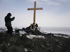 Personne chaudement habillée, de profil, à côté d'un croix en bois et en fond la banquise.