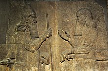 A stone relief showing Sargon II on the left wearing a crown and holding a staff facing a man on the right