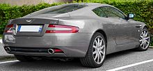 Rear three-quarters view of a bronze-coloured DB9 with its front wheel turned. There is a leafy bush in the background.