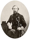 Sepia toned oval photo shows a bearded man with piercing eyes. He wears a dark military uniform and holds a sword with his gloved hands.