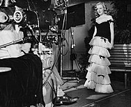 Monroe wearing a dress and facing a movie camera in a publicity photograph taken during her first film contract