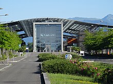 Photo sur l'entrée d'un bâtiment comprenant une grande porte vitrée et des toitures en acier.