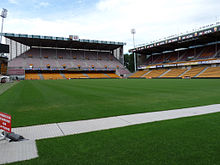 Photo d'un stade de football, la pelouse et deux tribunes sont visibles.