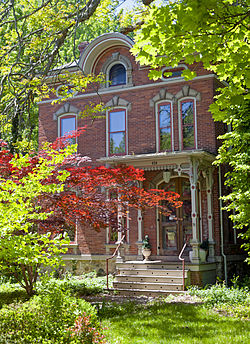 A brick house with ornate arches above its windows seen from slightly to its left though red and green leaves catching the sunlight