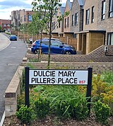 A white metal sign with black posts is shown in the foreground with the inscription "Dulcie Mary Pillers" in black lettering and the postcode "BS7" in red lettering. Houses constructed of red brick are shown in the background.