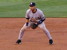 Baseball player in an athletic stance. He is wearing a blue baseball cap, and a grey jersey with the words "New York" inscribed.