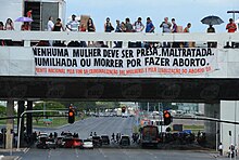 People stand on a highway with a banner hanging down that reads: Menhuma mulher deve ser presa, maltratada. humilhada ou morrer por fazer aborto. Prente nacional pelo fim da criminalizacao das mulheres e pela legalizacao do aborto.