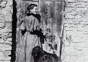 Photographie noir/blanc d’une femme, de dos, la tête tourné vers la droite, devant la porte d’un bâtiment vraisemblablement rural. À ses côtés, un chien noir.