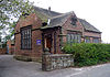 A single-storey brick building with a gable on the front, a door on the side and a short wall and a hedge in front.