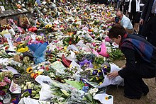 A woman adds a flower arrangement to a large memorial display set against a fence.