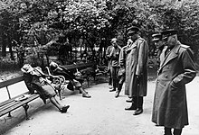 Six hommes en uniforme regardent trois cadavres reposant sur les bancs d'un jardin public. L'un de ces corps est couché, tandis que les deux autres sont assis, la tête basculée en arrière ou sur le côté.