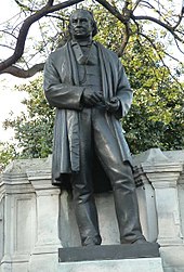 A bronze metal sculpture of a nineteenth century man wearing a long jacket or coat, trousers, waistcoat, with draughtsman's tools in his hands