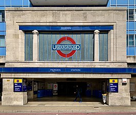 Station entrance in the form of a white stone-clad box sitting on two substantial and wide stone blocks. The front facade of the box contains a large London Underground logo (red ring with blue horizontal bar across the centre containing the word "UNDERGROUND") in the centre. Set back behind the entrance and to both sides a four-storey office block with blue cladding rises up.