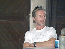 Jiří Langmajer seated, wearing a white Carhartt t-shirt, with arms crossed and sunglasses perched on his head, looking left of camera with a slight smile