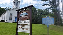 a picture of the marker with the white church and the church's own sign in the background