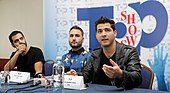 Three men at a conference sitting in front of a table with microphones. The man on the left is wearing a black shirt and has his hand holding his face. The man on the center wears a blue sweater. The man on the right wears a leather black jacket.