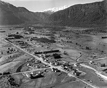 Black and white aerial photo of small West Coast crossroads settlement surrounded by forested mountains