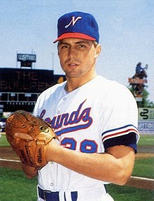 A man in a white baseball uniform and blue cap standing on a field with hands together in his brown leather glove