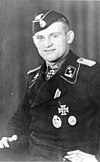 A man wearing a military uniform, side cap and neck order in the shape of a cross. His cap has an emblem in shape of a human skull and crossed bones.