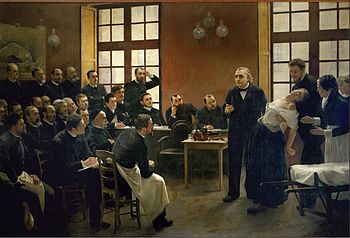 A painting of a 19th-century medical lecture. At the front of the class, a woman faints into the arms of a man standing behind her, as another woman, apparently a nurse, reaches to help. An older man, the professor, stands beside her and gestures as if making a point. Two dozen male students watch them.