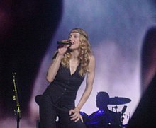 Madonna singing onstage wearing black pants and waist coat. She holds a microphone in her right hand to her mouth, while her left hand is supported on her thigh.